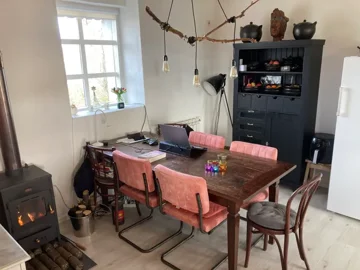 Dining area featuring a wooden table surrounded by pink chairs, a black cabinet with decorative items, a window, and hanging light bulbs from a branch.