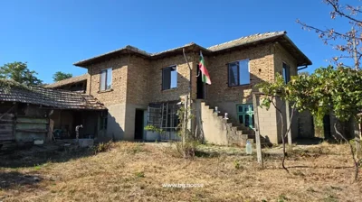 Two-storey brick house with tiled roof and external staircase, surrounded by dry grass and some trees under clear blue sky