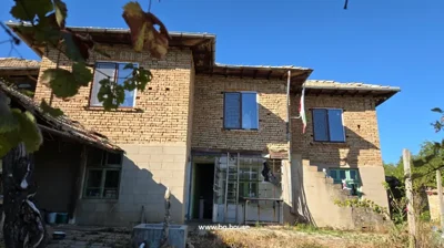 A two-storey brick house with green windows, a Bulgarian flag on a pole, and an open door with a glass panel in front.