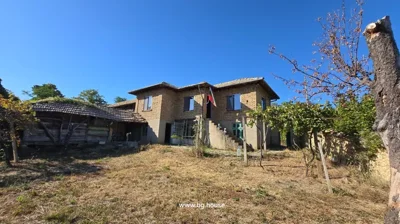 Wide view of two-storey brick house with tiled roof and external staircase, dry grass and trees in the garden under clear blue sky