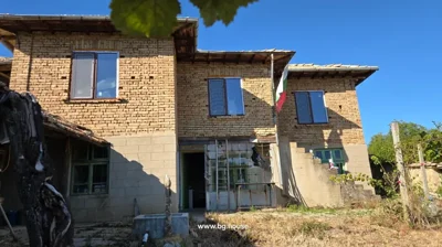 A two-storey brick house with green windows, a Bulgarian flag on a pole, and an open door with a glass panel in front.
