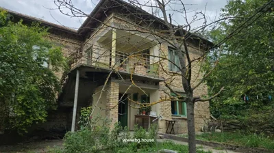 Two-storey brick house with green doors and windows, surrounded by trees and garden plants