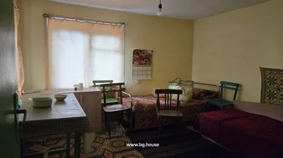 Room featuring two beds covered with patterned blankets, three wooden chairs, a table with a checkered tablecloth, a small cabinet, and a calendar on the yellow wall.