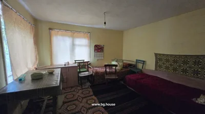 Room showing two beds with patterned blankets, three wooden chairs, a table with a checkered tablecloth, a small cabinet, and a calendar on the yellow wall.