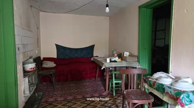Room featuring a bed with a patterned cover, a table with a checkered tablecloth, several chairs, and a stove on the left wall.