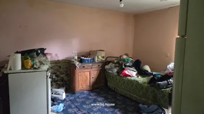 Room showing a bed covered with clothes, a wooden cabinet, a refrigerator, and various items scattered around.