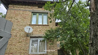 Side view of two-storey brick house with green window frames, satellite dish, and trees around.