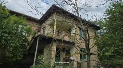 Two-storey brick house with green doors and windows, surrounded by trees and garden plants