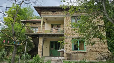 Front view of two-storey brick house with balcony, green doors and windows, and garden with trees