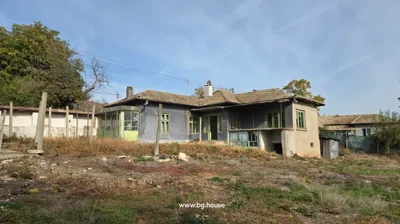 A single-storey house with a tiled roof and green window frames, surrounded by dry grass and trees under a clear blue sky.