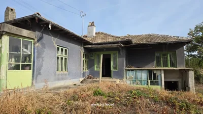 Front view of the house showing entrance door and windows with green frames, dry grass in front.